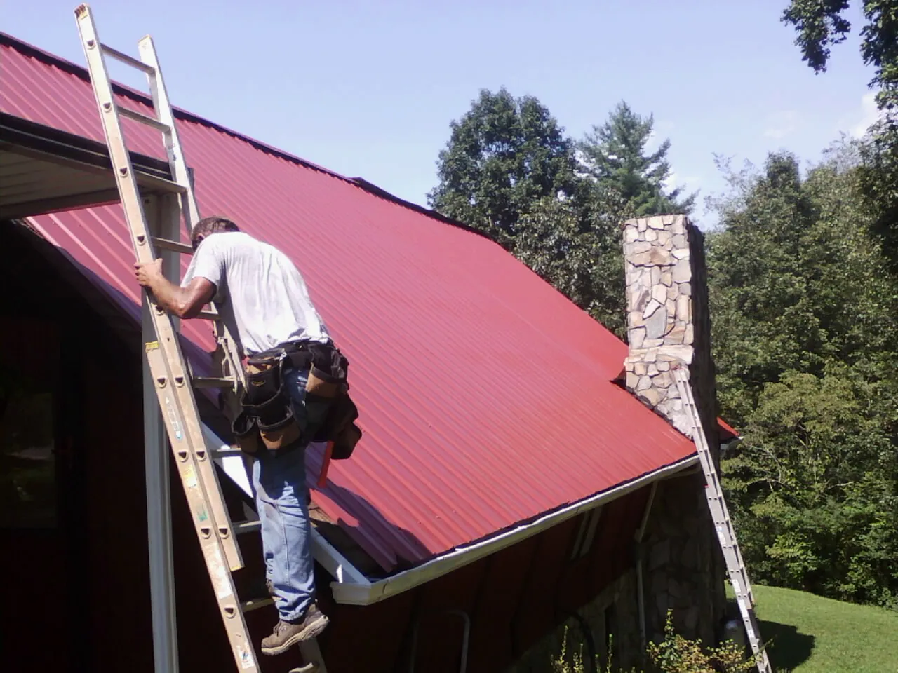 Roofer on a ladder beside a red metal roof and stone chimney — Soffit & Fascia Repair by Roofing Brothers in Granite Falls